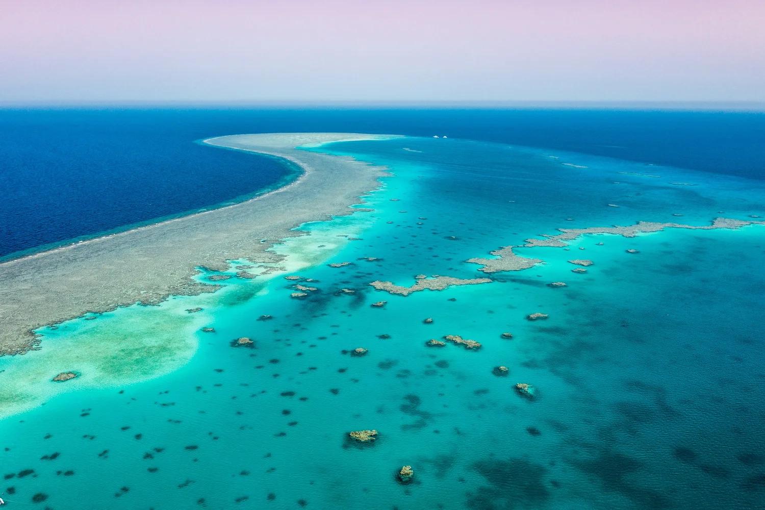 Aerial view of a curved coral reef with a turquoise lagoon and two boats on the horizon